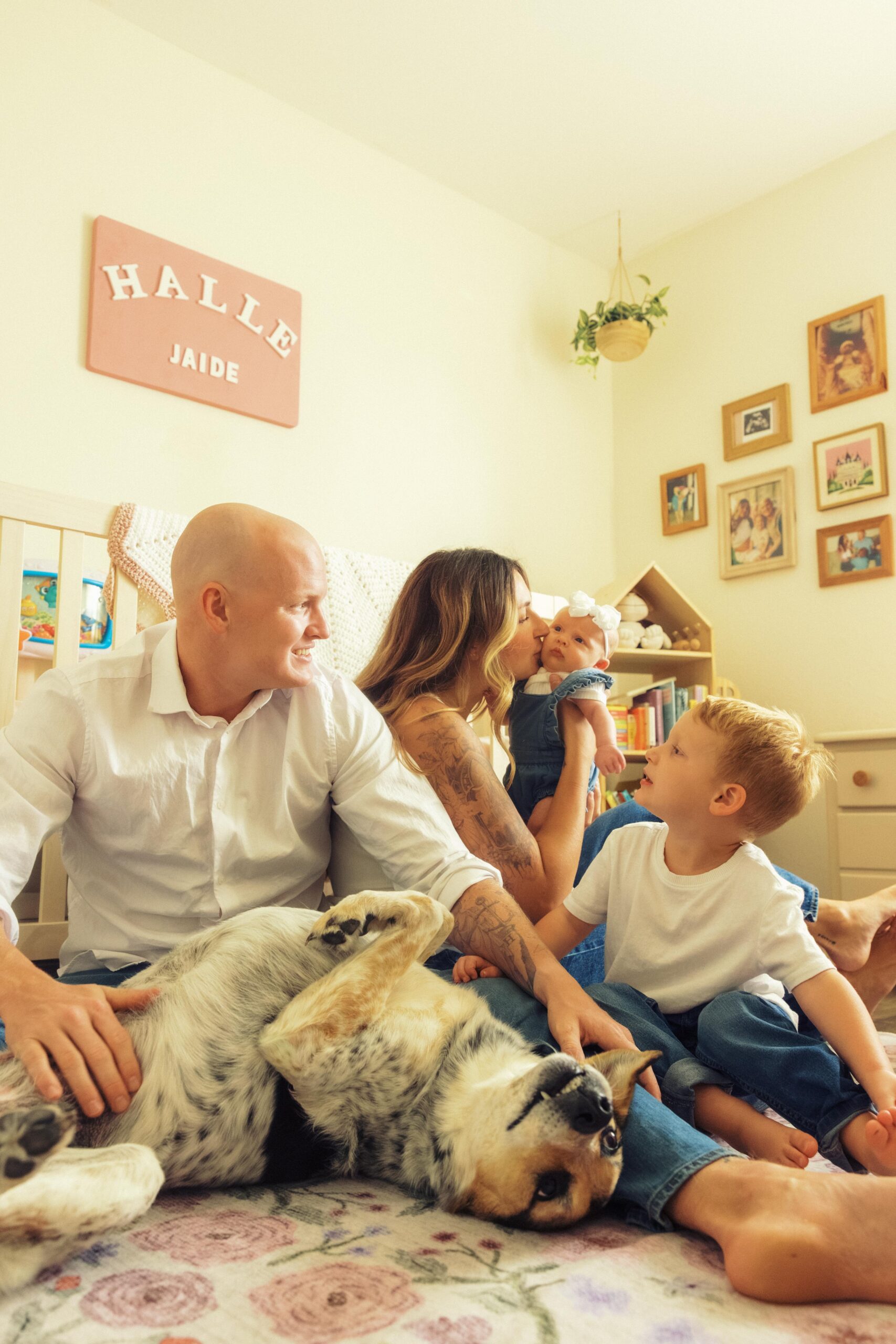 Lifestyle Newborn Nursery Photoshoot in Pearland TX with a family of 4 posing in front of the baby crib with their pet dog.